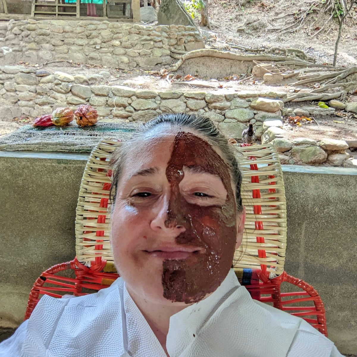 Misty in a chair outdoors, half her face covered in a cacao mask, with cacao pods on a wall behind her